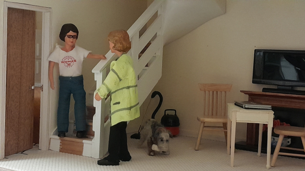 A 1:12 scale diorama of a living room. There is a brown fire door to the left that is slightly ajar revealing the kitchen. An open plan staircase painted white, but with bare woodwork on the treads where the carpet is missing, is to the left in the living room. There is a small table and two chairs in the middle of the room, there is a laptop on the table. Behind them is a brown wood fire surround with a black flatscreen TV on top.

Harriet, a Caucasian wom with dark hair, glasses, a white tee shirt and blue jeans is standing on the bottom step of the stairs talking to Daisy.

Daisy, a Caucasian woman with fair hair, a grubby high viz coat, and black trousers is standing next to the newel post talking with Harriet.

Under the stairs, next to the 'Henry' vacuum cleaner Monty Dog, Harriet's grey and black dog, is chewing on a bone.