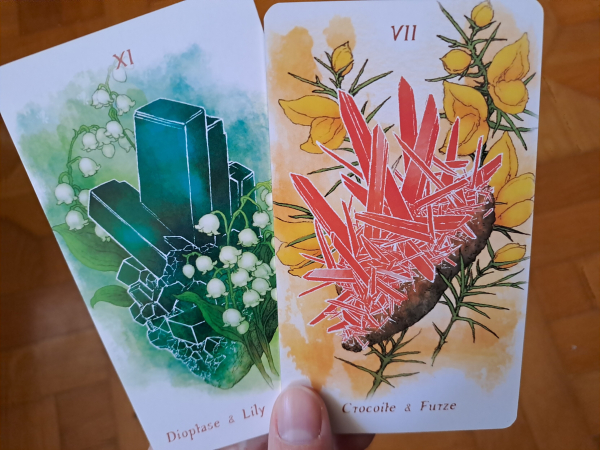 Photo of two colorful tarot cards. The one on the left features large green-blue dioptase crystals surrounded by lilies of the valley. The one on the right has spiky red crocoite crystals surrounded by yellow furze flowers.