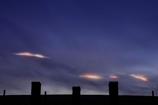 A dark silhouette of a roog top with three chimneys on and above the blue and a bit cloudy sky at dusk. There are four small stripes of polar stratospheric clouds near the roof. The one to the left, a bit higher up than the rest of them.