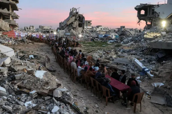 People gather by the rubble of destroyed buildings for a communal meal on a long row of tables snaking amongst ruins.