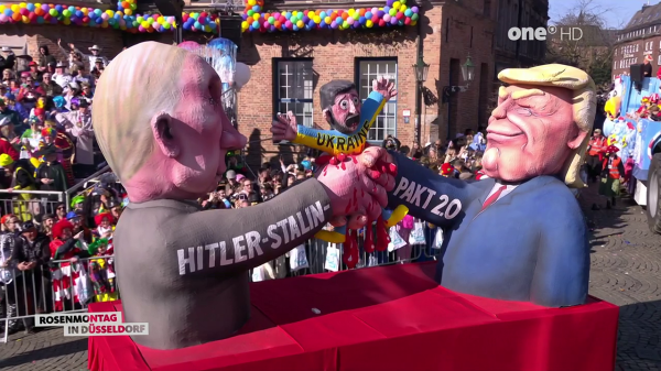 A full outdoor shot shows a parade float with large figures of Donald Trump and Hitler/Stalin shaking hands, with a figure of a person wearing the colors of the Ukrainian flag crushed in their hands. The hand shake is covered in what looks like blood. The Hitler/Stalin figure is wearing a gray shirt that says "Hitler-Stalin" while the Trump figure is wearing a blue suit with a red tie. The arms are labeled "Pakt 2.0". The crowd is blurred behind the float. The float is red. A logo for "Rosenmontag in Düsseldorf" is in the bottom left corner. A "one HD" logo is in the top right corner. A building is in the back.