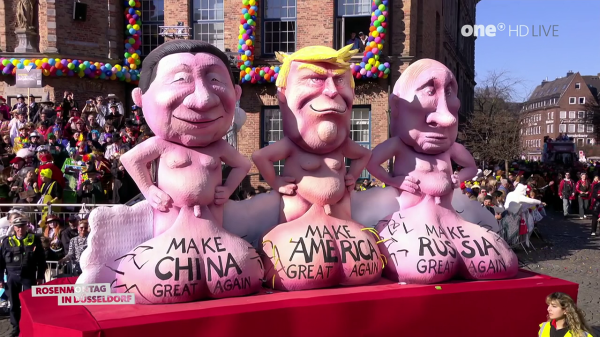  A full outdoor shot of a parade with a float of caricatures of world leaders. The caricatures are of Xi Jinping, Donald Trump, and Vladimir Putin, they are pink, topless, and pot-bellied, with their hands on their hips. Their lower bodies are shaped like testicles and have the phrases "Make China Great Again", "Make America Great Again", and "Make Russia Great Again" written on them. The float is red. Behind the float are crowds of people and buildings. In the top right corner of the image is the logo "one HD LIVE" and the words "ROSENMONTAG IN DUSSELDORF" are in the bottom left corner.