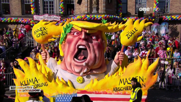 A wide shot shows a political float at a parade. The float is a caricature of Donald Trump, depicted with a red face, open mouth, and blonde hair styled into a laurel wreath. He is surrounded by yellow flames with German words written on them: "Annektionen" (annexations), "Zölle" (tariffs), "Klima-Störung" (climate disruption), and "Massenabschieben" (mass deportations). The float is decorated with the American flag and the text "Rosenmontag in Düsseldorf" (Rose Monday in Düsseldorf). Behind the float, a large crowd of people dressed in costumes watches the parade. A building with windows and a colorful balloon arch is in the background. The logo of "one HD" is visible in the upper right corner.