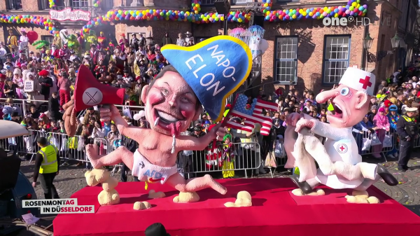 A wide shot shows a parade on a sunny day, with a crowd of people in the background, and two large cartoon figures on a red carpet. On the left, the first figure is a baby-like caricature wearing a blue "NAPO ELON" hat, holding a torn American flag in one hand and a red megaphone with a white "X" on it in the other. Its tongue is sticking out, and it's wearing a diaper labeled "AFD". The second figure, on the right, is a doctor-like caricature with a large nose, wearing a white coat with a red cross and a nurse hat with a red cross. It appears to be holding a straight jacket. In the lower left corner, a sign says "ROSENMONTAG in DÜSSELDORF".