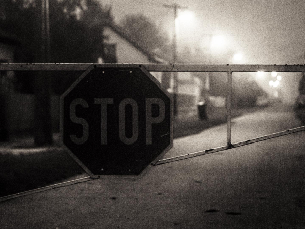 Straße mit einer Sperre plus Stoppschild vorn. Leichter Dunst, etwas vergangen. Eine Reihe Laternen. Schwarzweiß, körnig.