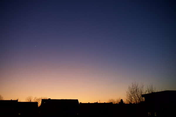 Der Abendhimmel mit Venus über einer Reihe dunkler Häuser.
Direkt über den Dächern ist der Himmel orange weiter nach oben violett bis blau.