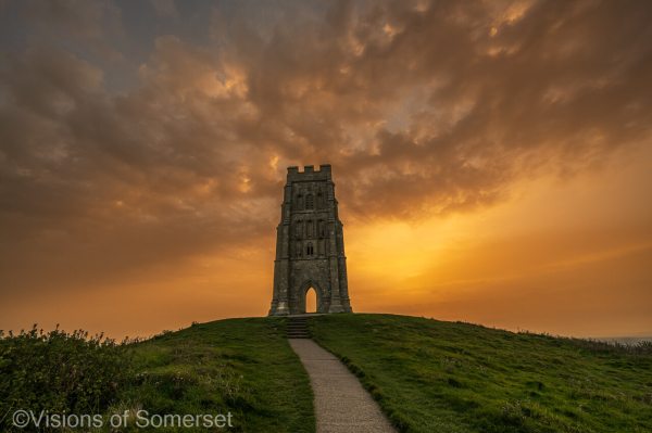 Golden sky and clouds behind the tower which is central in the scene. A pat leads up the hill.