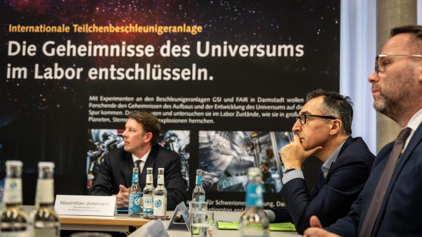 Minister Özdemir hört die Präsentation des Teilchenbeschleunigers, im Hintergrund steht eine Infotafel mit der Aufschrift "Die Geheimnisse des Universums im Labor entschlüsseln."