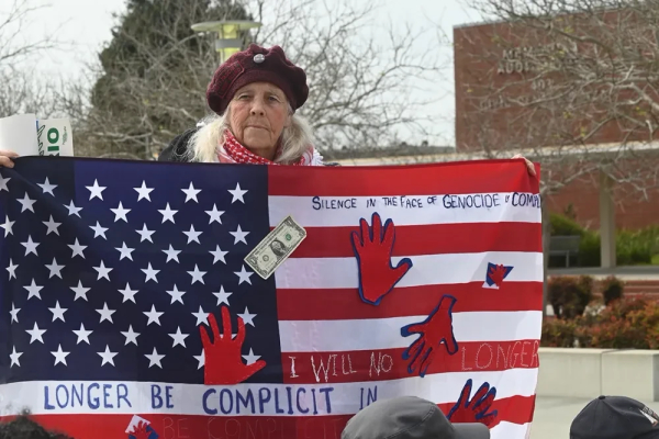 An older lady with shoulder-length white-grey hair, wearing a red beret, is holding a large placard of the American flag, with red hand prints on it.  It also has a dollar bill stapled to the middle.  Two phrases have been written on it:

SILENCE IN THE FACE OF GENOCIDE IS COMPLICITY

I WILL NO LONGER BE COMPLICIT IN GENOCIDE