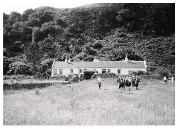 Black and white photograph showing a group of ramblers in front of a long white cottage beneath a wooded bluff.