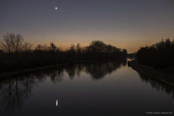 A calm river reflecting the fading light of dusk after sunset. The water's surface is smooth, mirroring the deepening hues of the evening sky. Above the distant tree line on the riverbank, the bright planet Venus shines prominently, its reflection shimmering on the water. Near the yellowish horizon, just above the trees, the faint glow of Mercury is also visible, adding to the serene and celestial atmosphere of the scene.