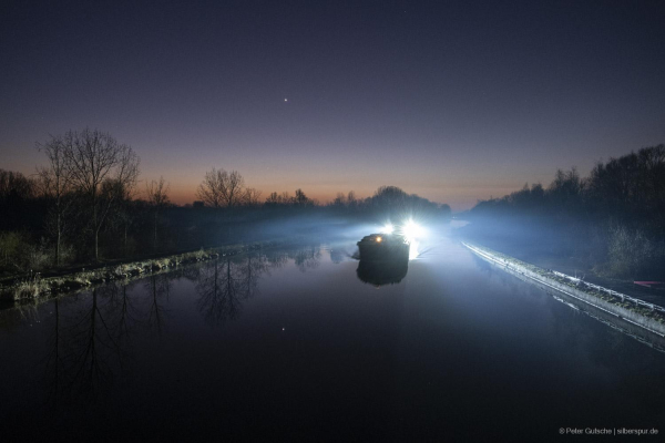 A calm river at dusk after sunset, its still surface reflecting the soft colors of the evening sky. The bright planet Venus shines prominently above, casting a shimmering reflection on the water. Near the yellowish horizon, just above the tree line on the riverbank, the faint glow of Mercury is visible. In the center of the image, a ship approaches on the river, its bright navigation lights shining directly toward the photographer. Despite the glare, the delicate presence of Mercury can still be seen in the sky.