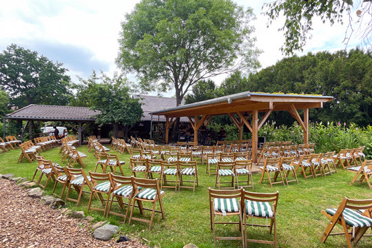 In einem großen sommerlichen Garten stehen in leicht gebogener Anordnung vor einer offeen Remise Holz-Klappstühle mit grün-weiß gestreiften Sitzpostern. 