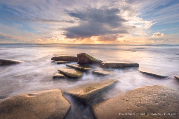 A seascape photo showing large, smooth rocks in the foreground, and a stormcloud out over the ocean. The sun is low in the sky and behind the stormcloud. Waves wash around the foreground rocks and the water's movement is rendered smooth and silky with a slow shutter speed. 