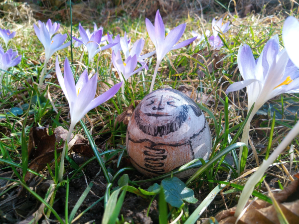 Inmitten grüner Grashalme und violetter Krokusse steht ein eiförmiges Stehaufmännchen. Es ist schwarzweiß bemalt und zeigt einen bärtigen Mann mit vor dem Bauch gefalteten Händen.