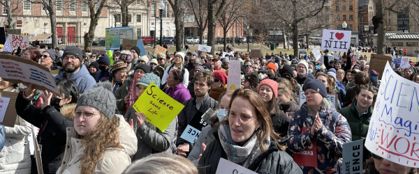 A crowd of mostly white people wearing winter clothing with signs such as "science saves lifes [sic]" and "Bad DOGE" and "I <heart> NIH"