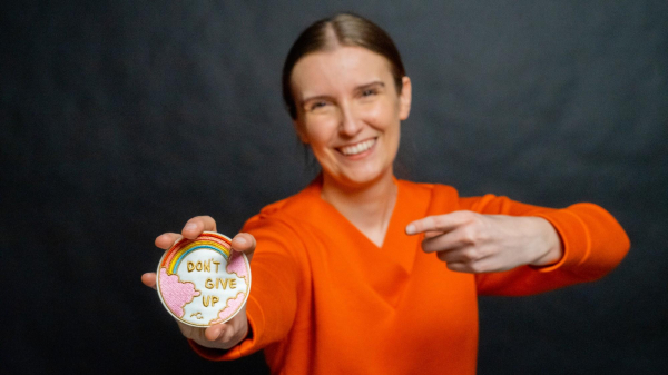 A smiling person in an orange sweater holds a round, colorful patch featuring a rainbow and the phrase "DON'T GIVE UP" on it. The background is dark.