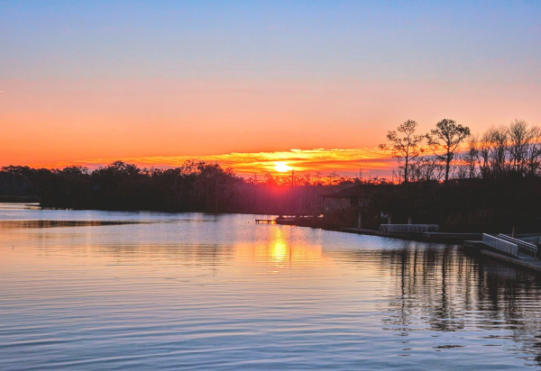 Warm glowing sunrise over a waterway. A waterway, lined with boat ramps, fishing piers, and darkened silhouettes of tree lines, reflects back a bright orange sunrise across the water's surface as the rising sun cast warm shades of pink, red, and orange across the morning sky.