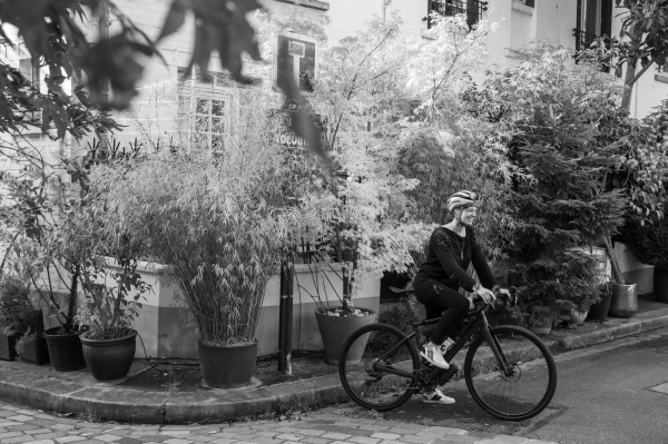 Photo en noir et blanc d'une cycliste prête à appuyer sur la pédale. De nombreux arbustes devant une maisonnette et une ruelle pavée.