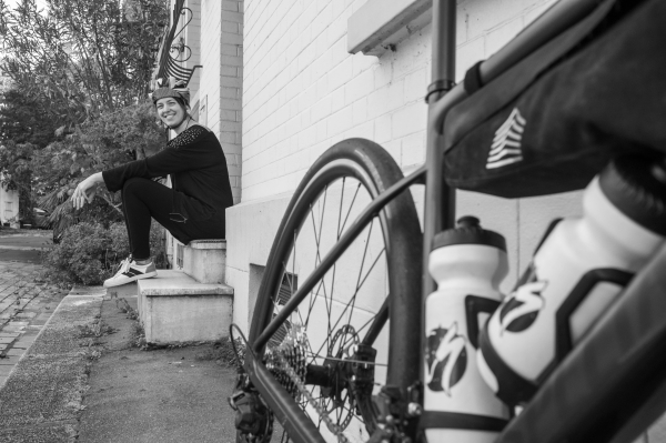 Photo d'un vélo en premier plan, on voit une partie du cadre et de la roue arrière. Derrière le vélo, une femme casquée assise sur le pallier d'une maison.