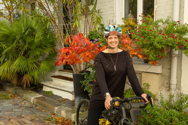 Photo en couleurs d'une cycliste sur son vélo. Elle porte une robe noir avec quelques sequins, et autour du coup un long collier en argent représentant un personnage à queue de cheval sur une roue de vélo.