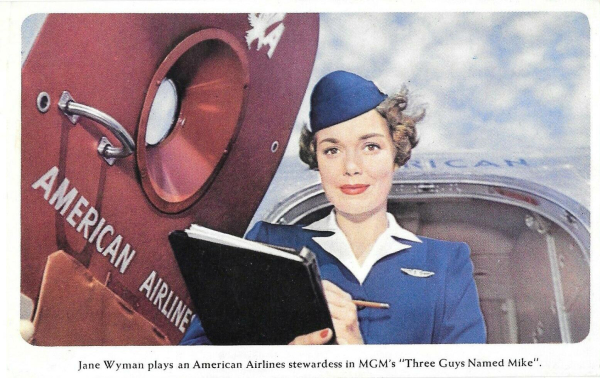 Jane is dressed as an American Airlines Flight Attendant and holds a a clipboard and pencil as she waits by a boarding door on a plane. 