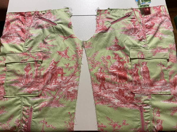 Two pants legs under construction facing each other at the crotch. Closeup view shows off how the cargo pockets match the underlying print from the front of each leg. Fabric is a pistachio green with magenta toile de Jouy (18th century French illustrations of rural life - this one is Asian rural life) print. Tableaus depicted are a boy on an elephant, a noble on a fancy litter, and two egrets grazing a marsh). Each tableau is approximately 12 inches wide and they have been centered on the high thigh area of the pants legs. 