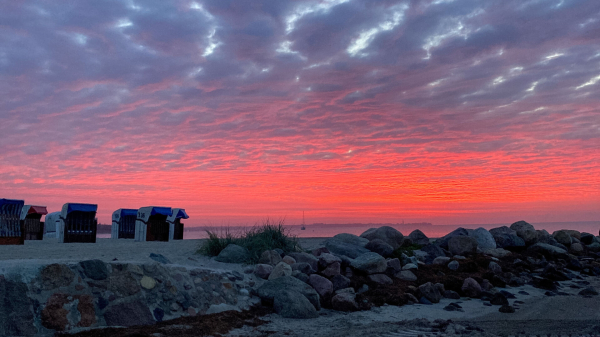 EIn mit dicken Wollen bedeckter Himmel leuchtet vom Horizont in knalligem Orangerot. Im Vordergrund Strand, abgesichert durch gestaplete Findlinge. Ein kleines Büschel Strandgras mittig hinter den Steinen. Links am Strand stehen. kreuz und quer 6 Strandkörbe. Zwischen Strand und Horizont erkennt man einen kleinen Steifen Ostsee auf dem ein Segelschiff (ohne Segel) fährt.