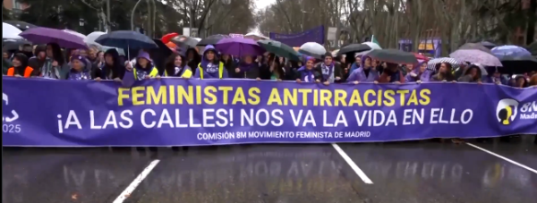 Huge crowd of protesters in a very wet Madrid marching behind a purple banner reading FEMINISTS, ANTIRACISTS, TO THE STREETS!  OUR LIVES DEPEND ON IT