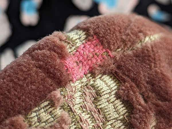 Close up on a hat made of a rose brown velour upholstery with floral woven into it. A rectangular patch of pink woven thread is up against a seam. 