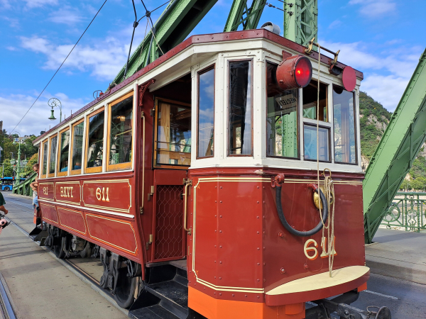 Photo of an old wood frame tram, painted brown, white and orange, with the number 611 on the front. Doors are a waist-high metal lattice that open and close by hand. In the background part of the green metal bridge structure is visible, and behind it Gellért Mountain.