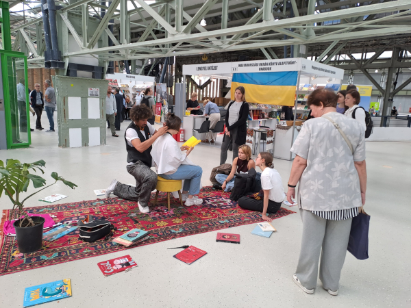 Photo from the book fair building. There is a colorful carpet on the floor, with books scattered on it. A girl sits on a small chair, reading from a yellow book, while a man kneels behind her, cutting her hair. There are people sitting and standing around, listening. 