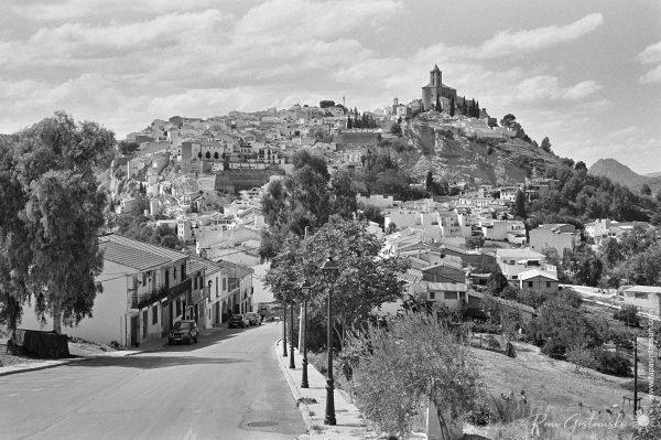 A black-and-white photo of the town of Iznájar on a hilltop. The prominent landmark is the Santiago Apóstol church.