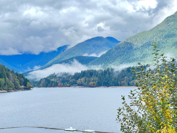 View of a lake with multiple mountains containing it. The mountains are mostly covered in green trees but there are a few trees with fall colors. It’s mostly cloudy but one of the mountains has been lit by sunshine. There’s a strip of dense fog going across the mountains.