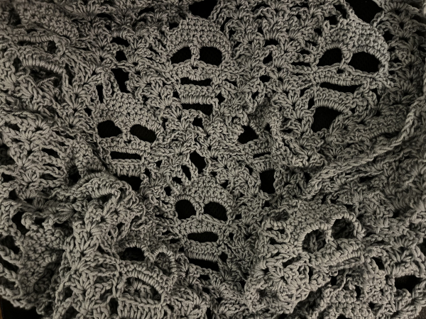 A grey crochet shawl with a pattern of skulls, on a black background. 