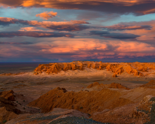 A distant view of red rock cliffs brightly illuminated in sunlight at sunset while foreground and background remain shaded by clouds. In the distance, clouds are illuminated as pink against fading blue.

